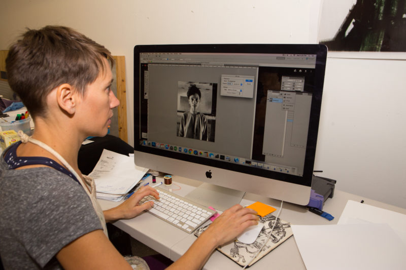 This is an image of a female student sitting in front of a computer. The computer screen shows a black and white photograph of a middle aged African American woman. The digital photo editing software Adobe Photoshop is open and shows multiple options for editing on the screen.