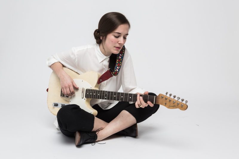 A woman sits on a white floor in front of a white wall with her legs crossed. She is wearing black capri pants and a white, button-down shirt. She has long, brown hair that is put into a bun-style by the back of her neck. She is holding a white electric guitar and looks to be strumming it.