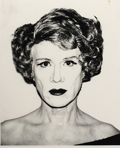 A black and white photograph shows Andy Warhol from the chest up in drag, wearing a short coiffed brown wig, heavy face powder, black eyeliner, and dark red lipstick. He stares at the camera, wearing a black shirt.
