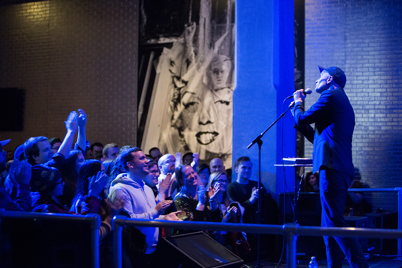 A man stands on a stage in The Andy Warhol Museum entrance space. He is singing into a microphone to a crowd of people in front of him.