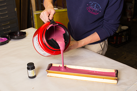 This is an image of a student pouring photo emulsion into a metal scoop coater. The photo emulsion is a bright pink and fills one half of the scoop coater.