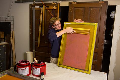 A student pulls a metal scoop coater filled with bright pink emulsion up the screen. The emulsion creates a big pink rectangle in the middle of the yellow screen.