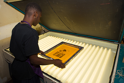 A student places a coated silkscreen on top of a film positive then onto a large light table. This light table has around twenty fluorescent bulbs and a black cover that goes over the table.