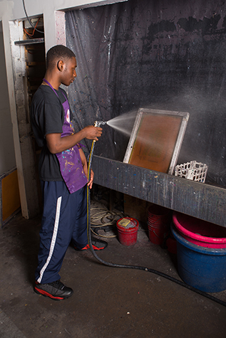 A student washes out a silkscreen using a hose, in a large sink. The sink has black plastic on the back and side walls.