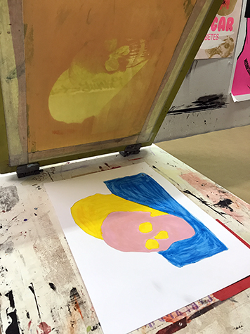 This image shows an underpainting of a skull being lined up and registered underneath a silkscreen with the same skull image exposed on it. The underpainted skull is painted pink, the shadow is yellow, and the ground is blue.