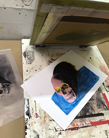 This image shows a final silkscreen print underneath a silkscreen that has been lifted up. The underpainted skull is painted pink, the shadow is yellow, and the ground is blue and the final photographic skull image is printed in black.
