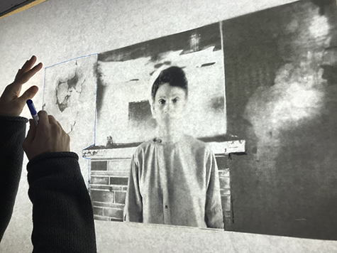 This image shows a set of hands to the left, using a blue marker to trace a square shape on a white piece of paper. Underneath the paper is a black and white photograph of a middle aged African American woman standing in front of a brick fireplace.