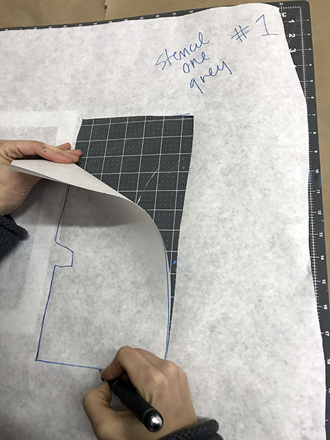 This image shows a set of hands cutting out a rectangle from a white piece of paper. The right hand cuts with an Xacto knife, while the left hand pulls the already cut piece from the paper. The words “Stencil one-grey” and the #1 appear in the upper right hand .