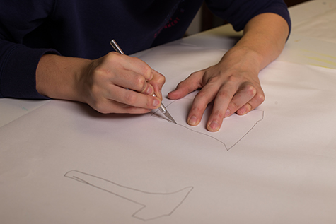 This image shows a pair of hands using an Xacto knife to cut out two outlined shapes from a white piece of paper.