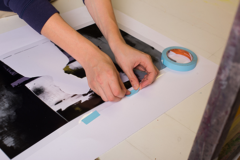 This image shows two hands using blue painters tape to secure a film positive to piece of printing paper.