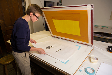 This image shows a male student with glasses placing a white paper stencil underneath a yellow and orange silkscreen to check for alignment. Two rolls of table sit on the table, one blue and one beige.