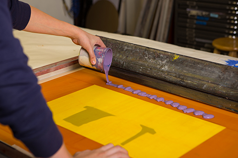 This image shows a hand pouring purple ink onto the top of a silkscreen. The silkscreen has two shapes cut out of paper underneath. A black squeegee rests at the top of the screen.