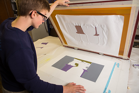This image shows a male student with glasses using a silkscreen and a paper stencil with two shapes cut out. The finished silkscreen print shows two shapes of color printed in purple on top of a grey background.