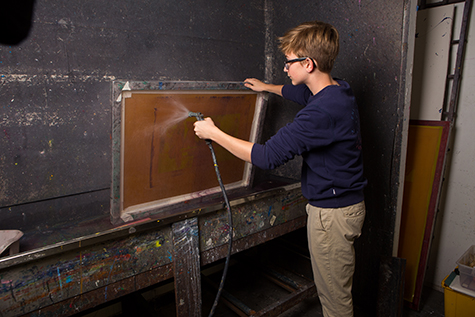 A student washes out a silkscreen using a hose, in a large sink. The sink has black plastic on the back and side walls.