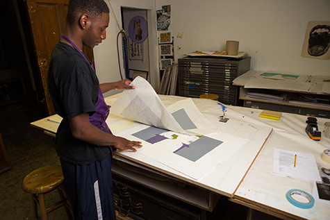 This image shows a student holding a stencil in his right hand. The stencil has been printed in grey ink and we can see two grey squares along with two purple and greenish-gold shapes on the white printed paper.