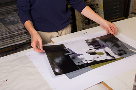 This image shows two hands holding a film positive over a multilayer print. We can see grey, purple and a greenish-gold showing through the film positive.