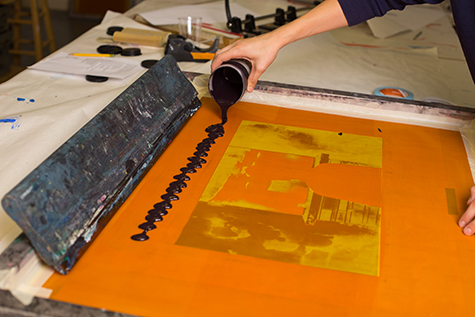 This image shows a hand pouring black ink onto the top of a silkscreen. There is a black squeegee resting at the top of the screen. We can see that an image has been exposed on the screen.