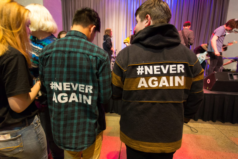 Two males are pictured with their backs towards the camera. The one on the left is wearing a green and black plaid shirt and the one on the right is wearing a brown coat with a hood. On their backs is the hashtag Never Again.