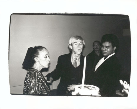A black and white photograph that has a woman standing on the left looking at a birthday cake being held by a man on the right. Andy Warhol is standing in the middle of the photograph blowing out the candle on the birthday cake.