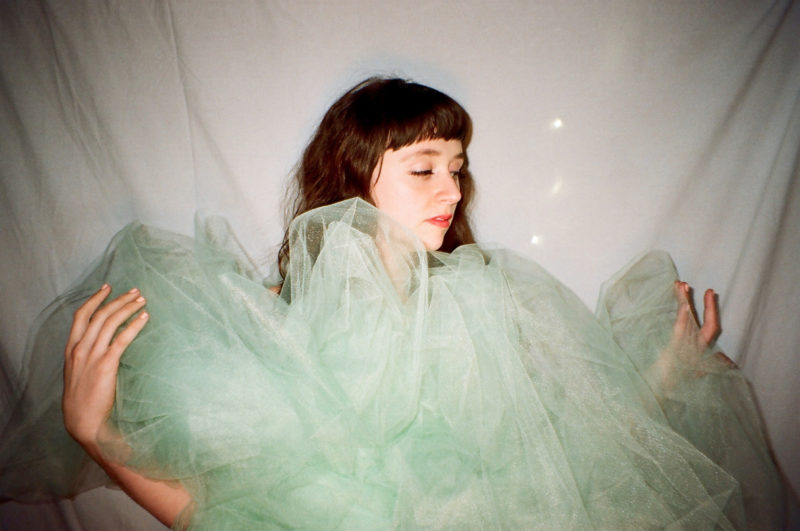 A woman stands in front of a white sheet. She has long, brown hair and it looking towards the right side of the frame. She is holding a a light green color sheer material that is bundled up to cover her body.