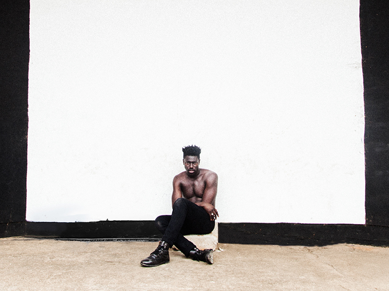 A shirtless man wearing black pants and boots sits on a white object in front of a white square painted on a black wall. He is looking directly at the camera.