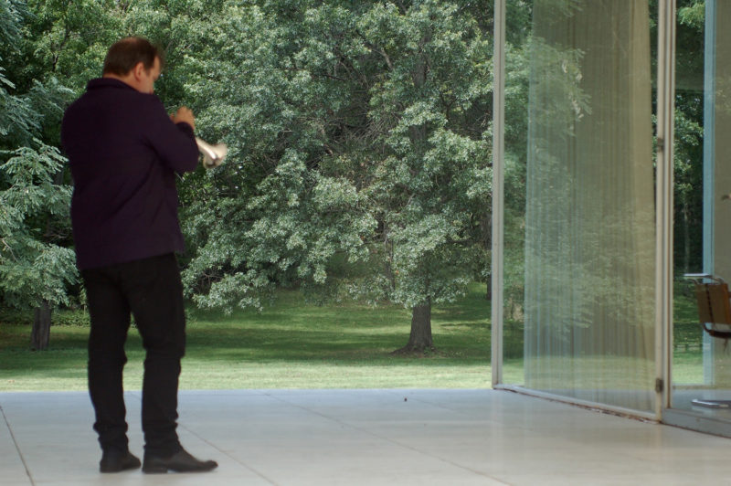 A man stands to the left of the image, with his back towards the camera. He is dressed in all black and appears to be playing the cornet, but only part of the instrument can be seen. He is facing the glass doors of a house, which has translucent blinds partially drawn. Dense trees can be seen in the background.