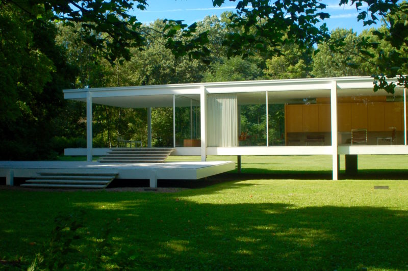 The image features a frontal view of the Farnsworth House. The crisp white, modern architecture contrasts the greenery surrounding the house. The white curtains are drawn, and mid-century furniture can be seen behind the windows that stretch from floor to ceiling.