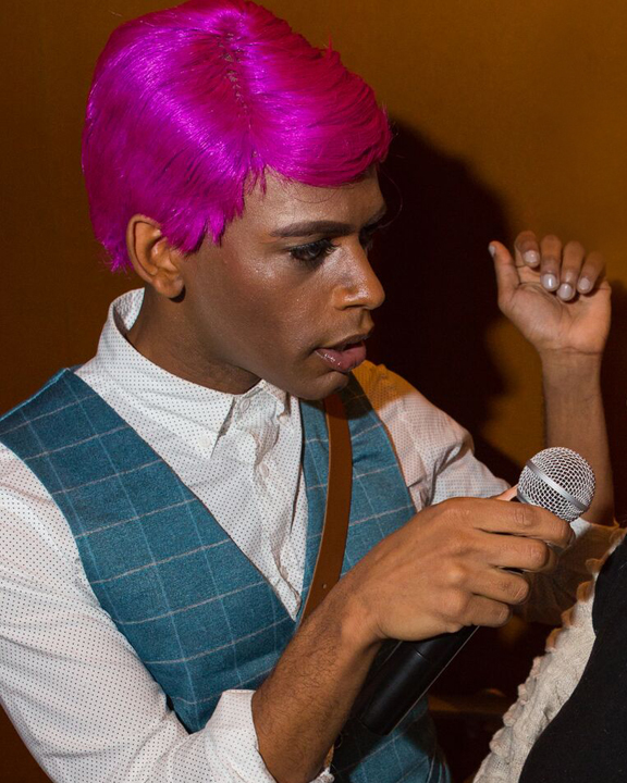 Artist Joseph Hall is wearing a hot pink, pixie-cut wig. He has on a white button-down shirt underneath a dusty blue vest. His right hand can be seen grasping a microphone in the bottom right corner of the image.