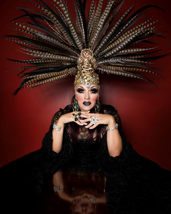 The image is a headshot of artist Mrs. Kisha Davis. The artist bears a giant, feathery headpiece that spans nearly half of the image. Their elbows are bent, leaning on a black tabletop, and their fingers are intertwined. The wall in background, is a deep, muted red.