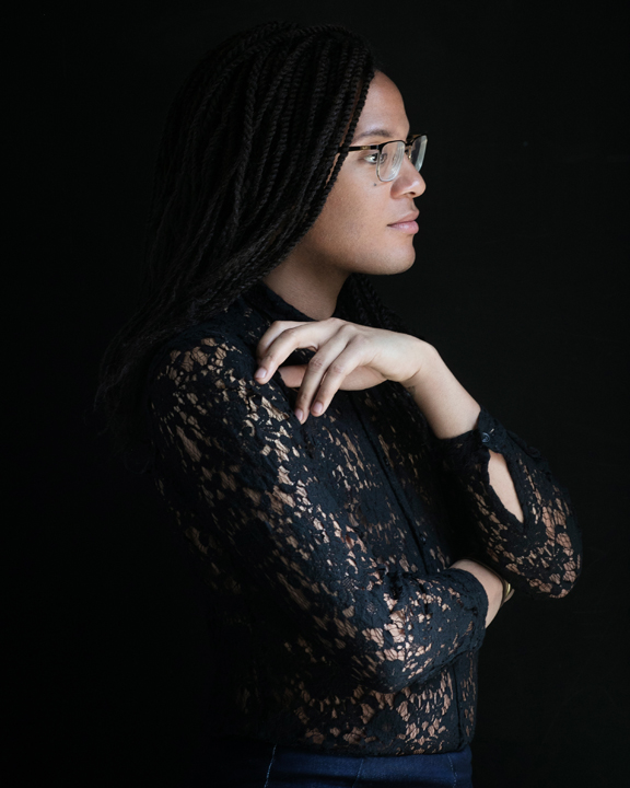 The image is a portrait of poet Ricky Laurentis. His profile is visible as he looks off to the right. He has glasses and long, braided hair that goes past his shoulders. His black lace shirt nearly blends into the black background. His left arm is bent, covering his torso, as his hand grazes his right shoulder.