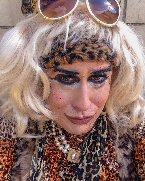 The image is a close-up of artist Scott Andrew. He is wearing a blond wig with white-framed sunglasses propped on top of his head. A fuzzy, cheetah print headband covers most of his forehead, and matches his cheetah print shirt and scarf. He is also wearing a chunky, pearl necklace bearing a gold pendant.