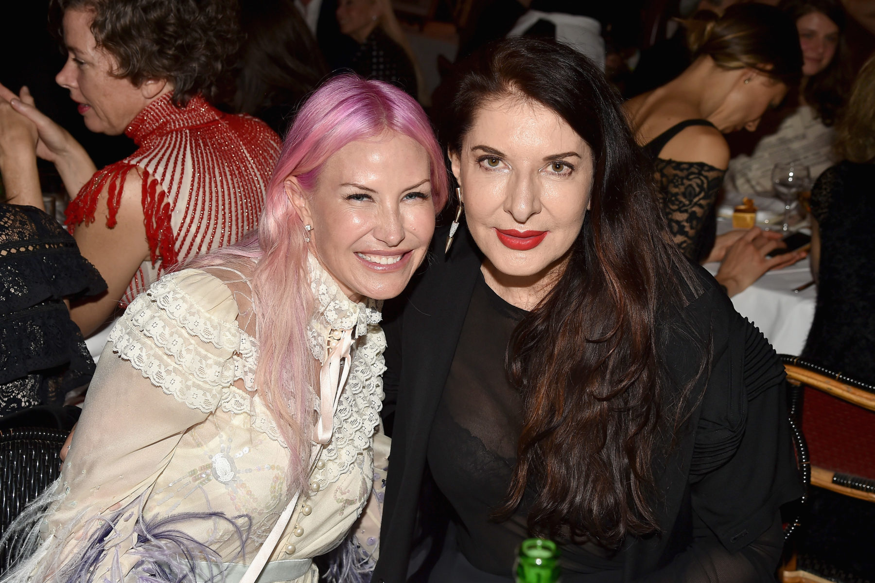 Two women sit at a table at the annual New York City dinner posing for a photo. The woman on the right has pastel pink hair and wears a white dress with lace and ribbon. The woman on the right is artist, Marina Abramovic. Marina has long dark brunette hair and wears a sheer black top and bright red lipstick.
