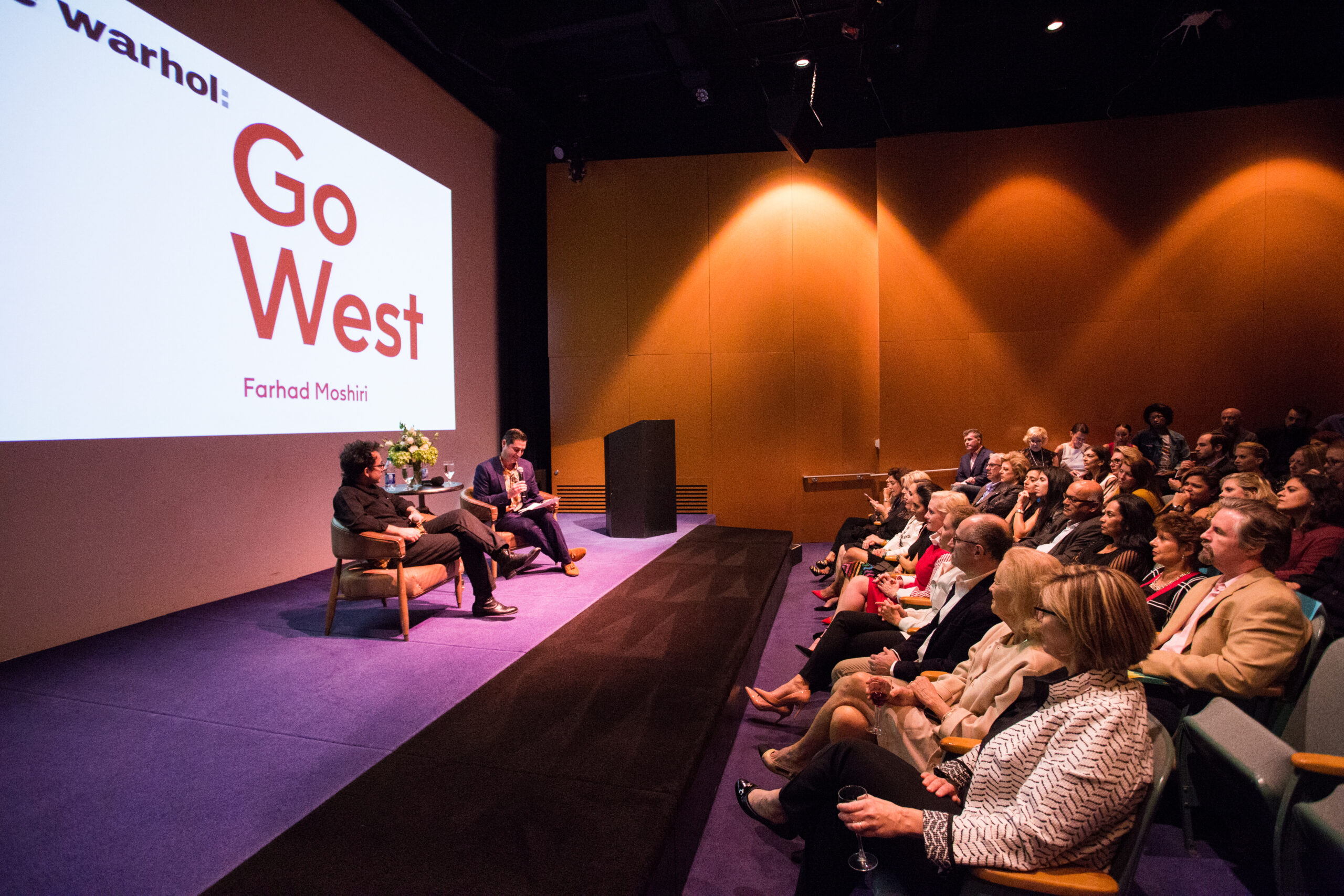 The camera is positioned at the far left of the stage panning out to the audience. The theater is filled with guests looking up towards the discussion occurring on stage. Two men, artist Farhad Moshiri and Chief Curator Jose Diaz, sit in discussion in front of a large projection that reads “Go West” in bold red font.