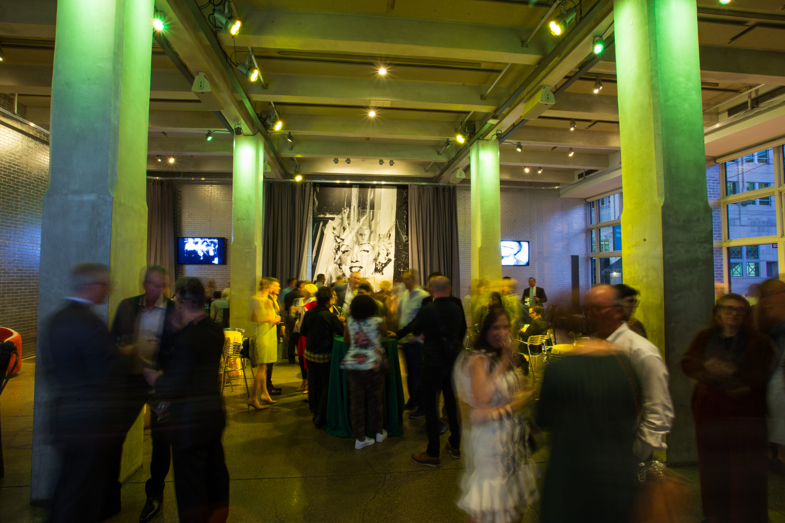 The Andy Warhol Museum’s lobby space is illuminated with bright green party lights as a crowd of patrons mingle. The image was taken using a long exposure making all of the moving people in the scene appear blurry as the architecture of the room appears clear.