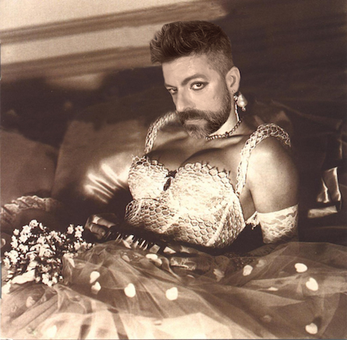 The sepia photograph features artist Miguel Gutierrez. They are leaning back, propped up by their elbows, with their head tilted towards the camera. Miguel is wearing a white lace, corset top and long tulle skirt. A bouquet of flowers lies on their lap.