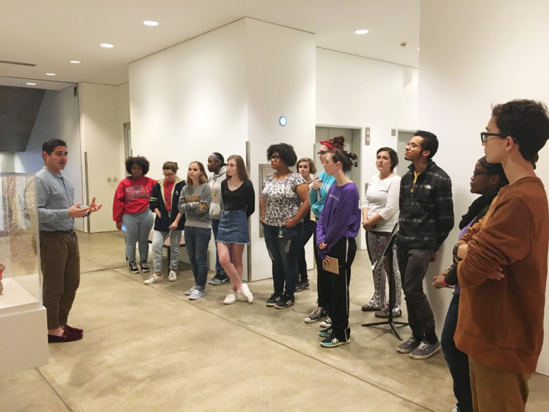 A large group of people are in a gallery listening to a man speak.