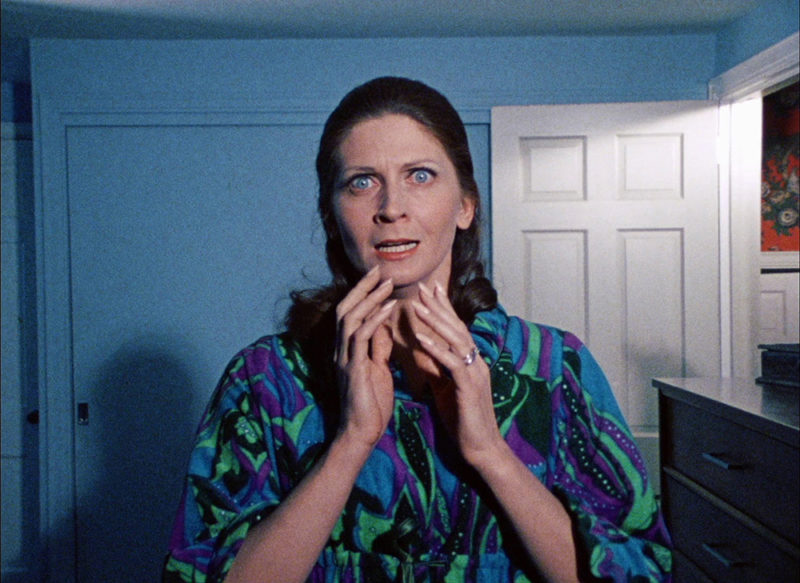 A film still depicting a woman looking towards the camera with her eyes wide open and her hands up towards her open mouth.