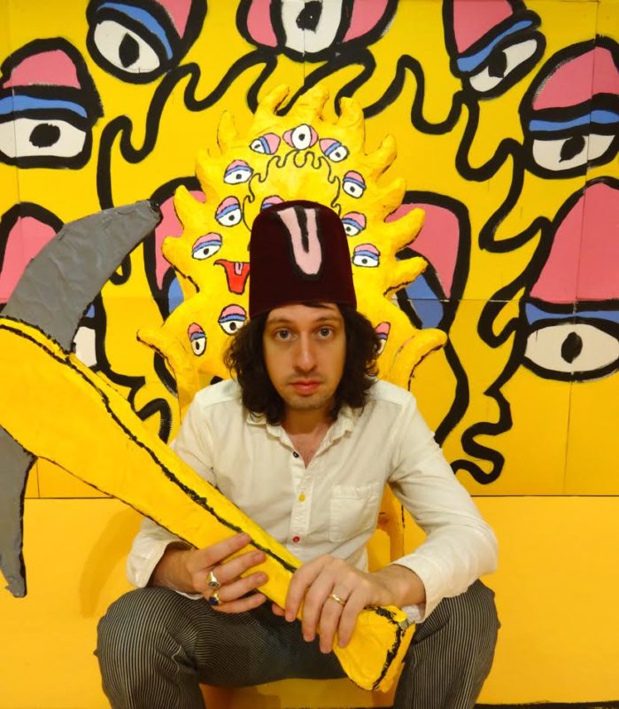 A man holding a cartoonish axe and wearing a tall, black hat is sitting in front of an artwork made of up painted cartoonish eyes on a yellow background.