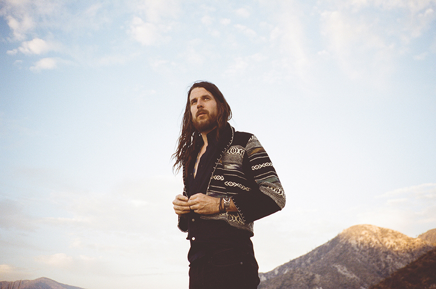 A photo of a standing man, taken from below his waist, pointing upwards towards his face. He is wearing black jeans and a black and white jacket. He has long, brown hair and brown facial hair. There are hills in the background.