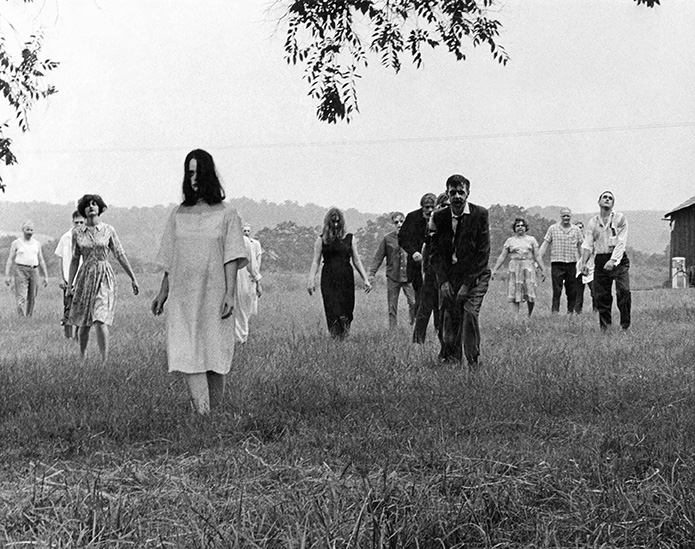 A black and white film still of male and female zombies in a field walking toward the camera.