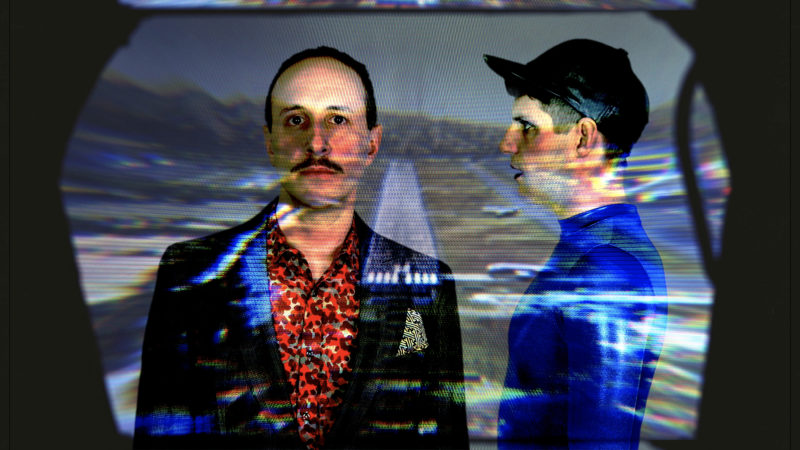 A photo of two men with an image projected onto them. The man on the left has a mustache and short, dark hair. He is wearing a suit coat over a multi-colored dress shirt, and is facing the camera. The man on the right is looking towards his left at the other man. He is wearing a black hat and blue shirt.