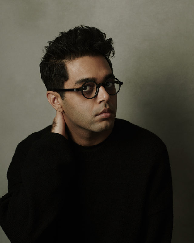 A man with short, dark hair and dark, round glasses looks at the camera. He is wearing a black, long-sleeve shirt and his right hand is on the back of his neck.