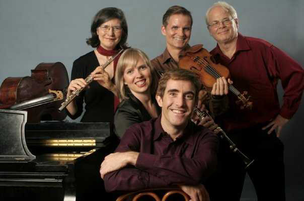 A group of two women and three men are smiling at the camera next to a cello and a piano. One woman holds a flute, the other holds a clarinet. One man holds a violin.