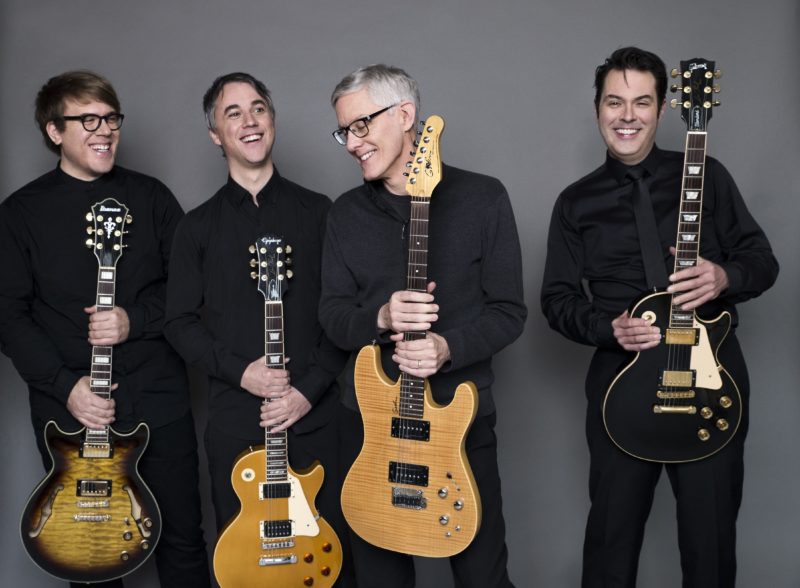 Four men in all black stand in a row while looking off camera and smiling. Each of them hold electric guitars by the neck.