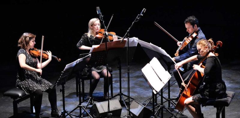 Three women and one man sit in a semi-circle on a black box stage playing stringed instruments. From left to right, a woman plays the violin, another woman plays the violin, a man plays the viola, and a woman plays the cello.