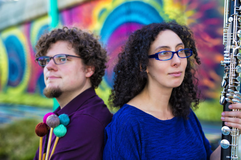 In front of a colorful mural, a man with glasses and a goatee holding marimba mallets stands back to back with a woman with glasses and curly hair holding a bass clarinet.