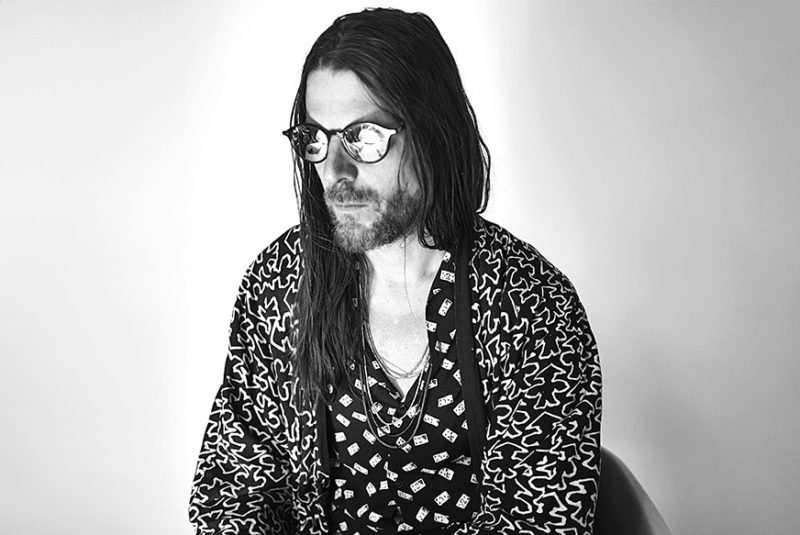 A black and white photograph of a man with long, dark hair and a mustache and beard, sitting on a chair. He is wearing glasses, necklaces, and a black and white patterned shirt, and looking off to the left of the frame.