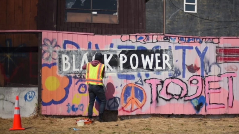 Person in a reflective safety vest paints “Black Power” in black and white onto a pink, graffitied wall.