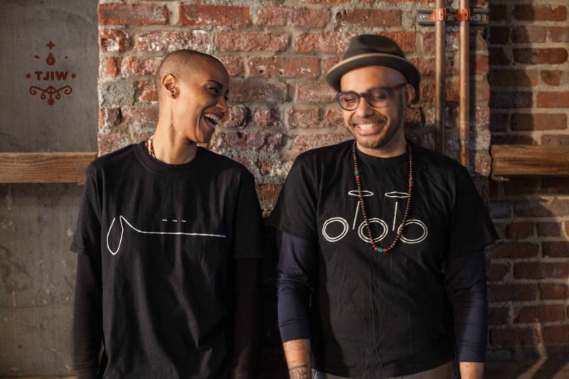 A woman with a shaved head and a man in a hat and glasses stand next to each other in front of exposed brick. They are both in black. She is looking at him and laughing while he is looking down and smiling.