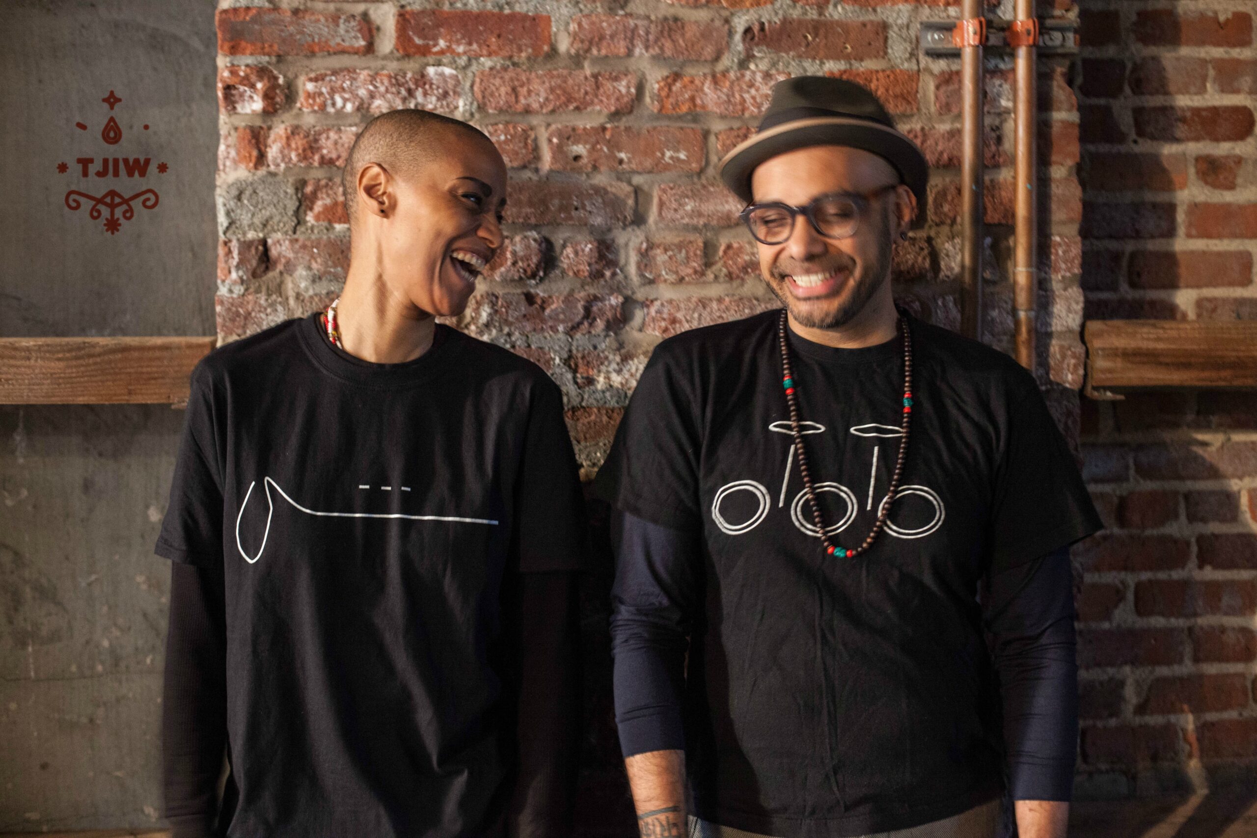 A woman with a shaved head and a man in a hat and glasses stand next to each other in front of exposed brick. They are both in black. She is looking at him and laughing while he is looking down and smiling.